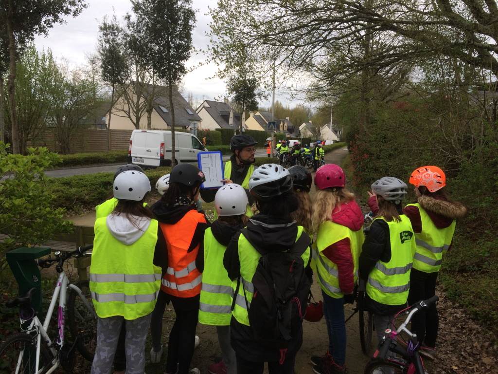 Un groupe d'enfants de dos avec des gilets et casque de vélo regardent avec attention les explication d'un cycliste adulte